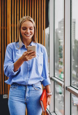 Neşeli kadın blogcu, ücretsiz 4G internetini kullanarak sosyal ağlarda modern akıllı telefon cihazında komik bildirimler okuyor. Ofis içinde cep telefonuyla sohbet eden olumlu genç kadın.