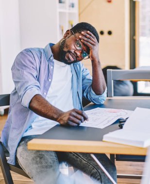 Yorgun sakallı siyah adam alnına dokunuyor ve masa başında oturmuş, ofis planları üzerinde çalışırken kameraya bakıyor.
