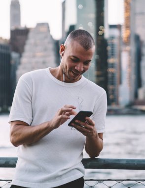 Genç gülümseyen etnik adam kulaklıkla müzik dinlerken akıllı telefon kullanıyor ve nehir ve şehrin bulanık arka planına karşı korkulukların yanında duruyor.