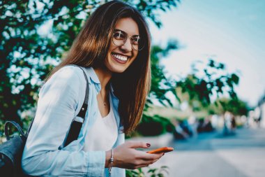 Sırt çantalı neşeli genç bir kadın, şehir ortamında gülüyor, elinde modern akıllı telefon tutuyordu. Günlük kıyafetler giymiş pozitif hippi kız, kamuya açık internet bağlantısını kullanarak cihaza güncelleme uygulaması yüklüyor.