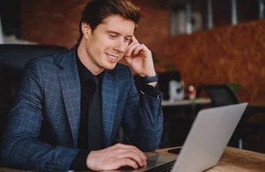 Kafkasyalı kendine güvenen erkek girişimci internet sayfasından gelen haberlerden memnun, netbook 'ta gezen gülümseyen iş adamı, netbook' taki uygulamayı kullanarak ofisindeki masaüstünde oturan muhasebe kayıtlarını kontrol ediyor.