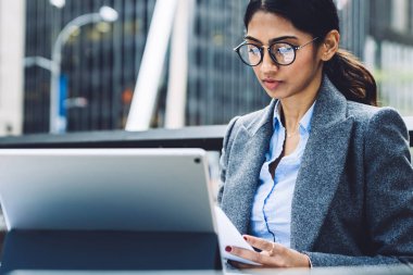 Kendine güvenen, gri ceketli ve gözlüklü iş kadını New York şehrinin terasındaki tableti incelerken şehir merkezindeki masada oturmuş belgeleri kontrol ediyor.