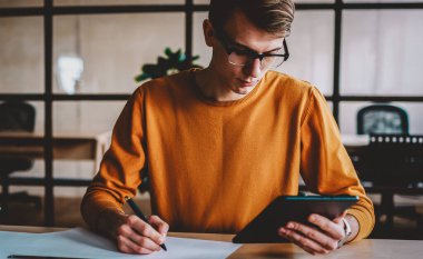 Modern tableti elinde tutan konsantre yaratıcı mimar ve stüdyoda ekipman ve tüple masaüstünde oturan bir binanın taslağını çiziyor. Düşünceli hippi adam plan üzerine not yazıyor.