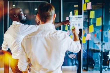 Başarılı projenin işbirliği sırasında cam duvara yapıştırılmış notlarla renkli etiketleri işaret eden Afrikalı Amerikalı CEO. Gururlu CEO ve modern ofis çalışanları arasındaki işbirliği.