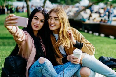 İlkbaharda yeşil çimenlerde oturup kahve içerek selfie çeken çekici bayan öğrenciler, şehir parkında mola vermiş modern akıllı telefonlardan fotoğraf çeken kadın blogcular.