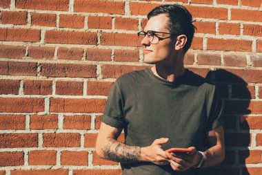 Gözlüklü, sıradan giyinmiş, cep telefonuyla mesajlaşan, sokakta duvara yaslanan ciddi bir genç erkek öğrenci.