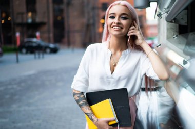 Sokakta cep telefonuyla konuşurken elinde not defterleri olan ve kameraya bakan aktif ve modaya uygun genç bir bayan.