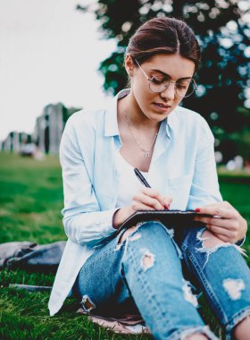 Zeki hippi öğrenci yeşil çimlerde oturuyor ve boş zamanlarında not defterinde ödev yapıyor. Sıradan giyinen zeki genç kadın parkta açık havada albümde resim çiziyor.