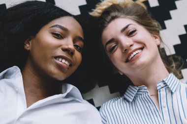 Beyaz bir arkadaşıyla kameraya gülümseyen Afro-Amerikan hipster kızı. Evde yerde yatan ve birlikte zaman geçiren iki çok kültürlü genç kadının portresi.