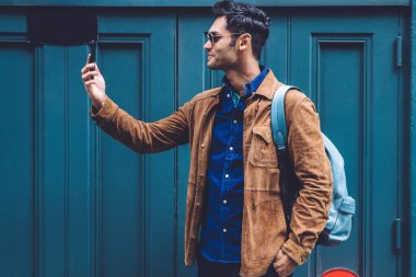 Sırt çantalı, modaya uygun giysileri ve güneş gözlükleriyle gülümseyen genç adam New York caddesinin mavi binasında dururken selfie çekiyor.