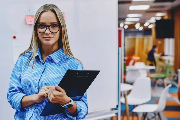 Portrait of charming young woman in eyeglasses working coach holding folder and standing against flip chat in classroom.Attractive female teacher looking at camera before workshop in modern office