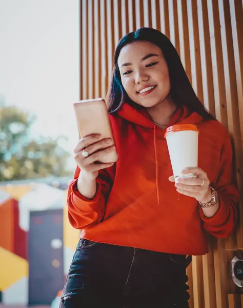 Concept of mobility and millennial time for generation, positive japanese female teenager with tasty takeaway coffee in hand reading news on funny content website during leisure in city environment