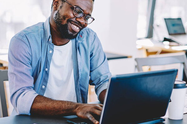 African American man in glasses working at laptop and smiling happily while sitting at table against large windows and desktops