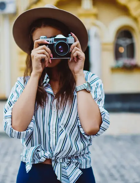 Sokakta fotoğraf çekmek için klasik kamera kullanan modaya uygun şapka ve giysili genç bir kadın, açık havada kentsel ortamların resmini yapan profesyonel bir kadın fotoğrafçı.