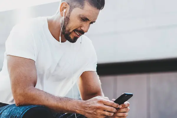 Bearded Caucasian male jogger dressed in stylish sportswear reading received message about success in cardio training connected to 4g wireless on mobile phone, concept of healthy lifestyle