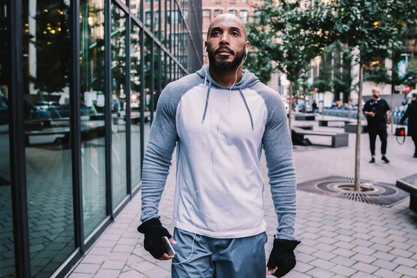 Strong African American man with earphones in sportswear walking and looking away during workout on street near modern office building