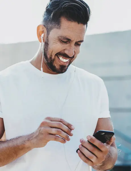 Positive mature man in electronic earphones watching funny video and laughing during leisure time on urban setting, happy male blogger enjoying online communication with followers from social website