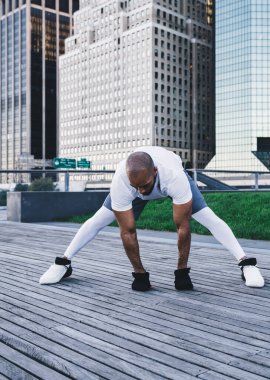 Spor kıyafetleri içinde güçlü Afrika kökenli Amerikalı bir erkek sabahları gökdelenlerin yakınındaki kaldırımda bacaklarına yoğunlaşarak diğer tarafa bakıyor.