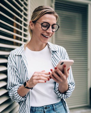 Neşeli, genç, güzel bir bayan günlük kıyafetler ve moda gözlükler takıp gülüyor, akıllı telefonlara bakıyor ve bina yakınlarında komik videolar seyrediyor.