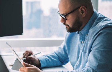 Bilgisayarda bilgisayarın başında dizüstü bilgisayarla otururken grafik tablet üzerinde çalışan odaklanmış sakallı adamın ekin kenarı görüntüsü