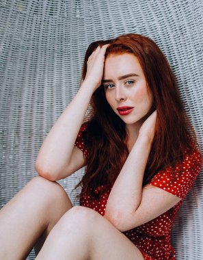 Gülümseyen, çekici, kızıl elbiseli, beyaz sallanan sandalyede dinlenen yeşil tropikal bitkilerin arka planındaki kameraya bakan genç bir kadın.