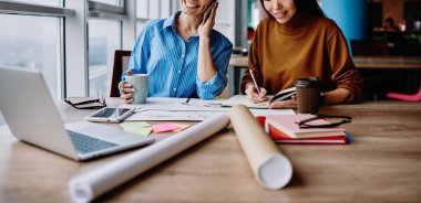 Masaüstünde birlikte tasarım yapmayı planlayan başarılı kadın ofis çalışanları. Akıllı telefondan arayan beyaz bir kadın. Neşeli meslektaşı not defterine bilgi kaydediyor.