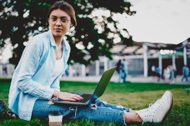 Ulusal parktaki yeşil çimlerde dizüstü bilgisayarla otururken kameraya bakarken sıradan giyinen genç bir kadının portresi. Netbook 'ta internet ortamında çalışan çekici hipster öğrenci.