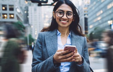 Yuvarlak gözlüklü, esmer, şık, genç bir kadın. Siyah el çantası ve gri ceketli. New York sokaklarında yaya geçidinde akıllı telefona dokunuyor ve kameraya bakıyor.