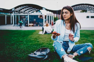 Modern akıllı telefon cihazındaki uygulamayı, modern parktaki yeşil çimlerin üzerinde oturan yüksek hızlı 4G internet bağlantısı üzerinden güncellerken, gündelik kıyafetlerini giymiş çekici genç bir kadın gözlerini kaçırıyor.