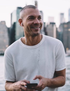 Beyaz tişörtlü, gülümseyen, kulaklıkla müzik dinleyen ve nehir kenarındaki metal tellere yaslanmış akıllı telefon tutan genç bir adam.