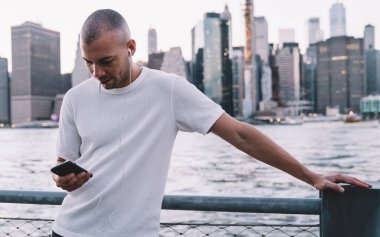 Beyaz gömlekli, gülümseyen, kulaklıkla müzik dinleyen ve nehir ve şehir yakınlarındaki metal çitlere yaslanarak akıllı telefon kullanan genç bir adam.