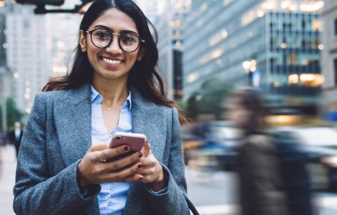 Yuvarlak gözlüklü, esmer, şık, genç bir kadın. Siyah el çantası ve gri ceketli. New York sokaklarında yaya geçidinde akıllı telefona dokunuyor ve kameraya bakıyor.