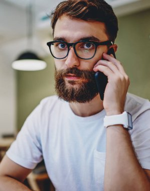 Ciddi bir beyaz sakallı adamın portresi cep telefonuyla bankacılığı arıyor içeride oturuyor, görüş düzeltmesi için gözlüklü genç bir hippi kameraya bakıyor telefonda konuşuyor.