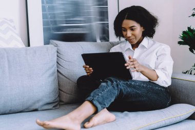 Düşünceli Afro-Amerikalı genç bir kadın internet sitesinden canlı yayın izliyor dijital tablette ev interneti kullanıyor rahat bir dairede koltukta dinleniyor. Yoğunlaştırılmış koyu tenli kadın haberleri okuyor.
