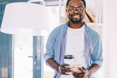 Neşeli, kendine güvenen, sakallı, gözlüklü, sıradan giysileri olan, hafif ofisteyken içecek ve akıllı telefon taşıyan siyahi adam.