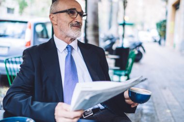 Caddedeki kafede gri sakallı, mavi kravatlı, kahve fincanı ve şehir arka planında gazetesi olan neşeli, son sınıf öğrencisi.