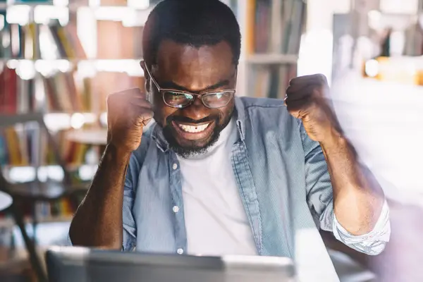 High angle of African American adult overjoyed male in glasses and casual clothes with arms raised celebrating victory while sitting at table and using laptop in light modern office