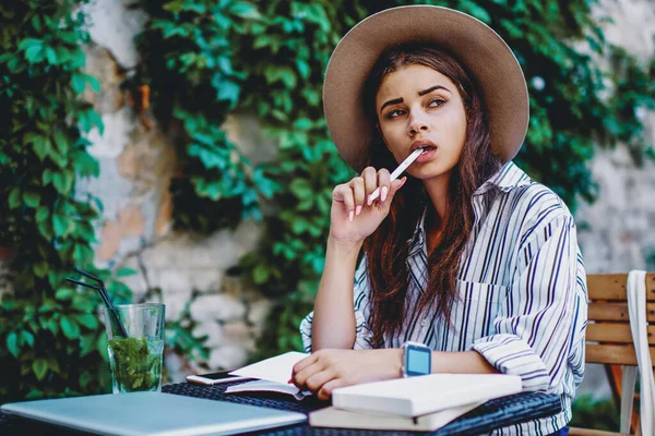 Pensive hipster girl in trendy hat pondering while creating article for blog holding pen in mouth sitting outdoors in cafe, brunette caucasian woman author writing in notebook making to do list