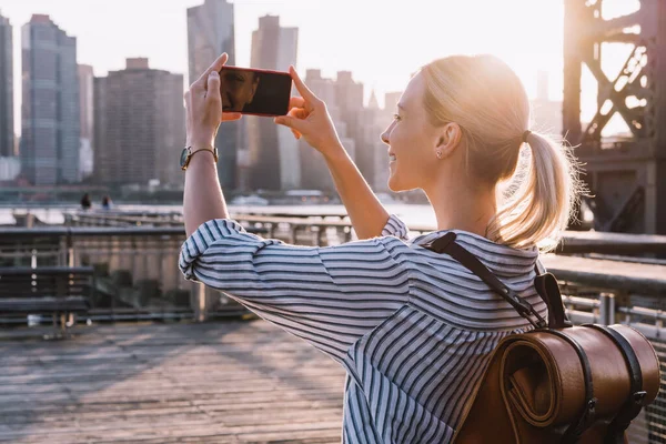 Back view of millennial woman shooting video of New York landscape using smartphone for clicking pictures, positive female tourist testing cellphone camera for taking photos of city in sunrise