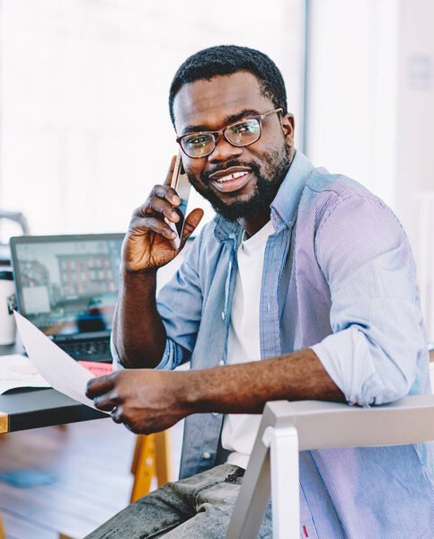 Smiling cheerful African American male talking on mobile phone holding document in hand sitting at table with laptop in office