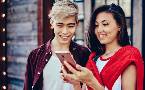 Positive chinese young man and woman reading funny notification in social networks on modern smartphone standing in urban setting.Cheerful hipster bloggers laughing while watching video on mobile