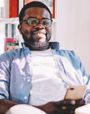Gözlüklü, rahat giyinen, sandalyede otururken kameraya gülümseyen ve akıllı telefon kullanan Afrikalı Amerikalı bir erkek.