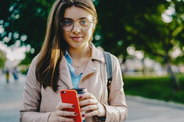 Moda gözlüklü, elinde kahve, elinde akıllı telefon olan çekici bir kadının portresi şehir ortamında caddede duruyor. Kafein içeceği olan hippi kız.
