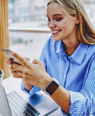 Ofisteki dizüstü bilgisayarında oturan dijital akıllı telefon cihazına yazı yazan zeki genç bir kadın. 4G kullanarak sosyal ağlarda medyayı paylaşan pozitif hipster kız.