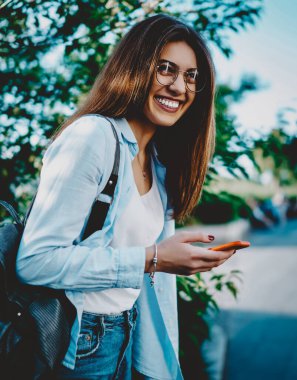 Sırt çantalı neşeli genç bir kadın, şehir ortamında gülüyor, elinde modern akıllı telefon tutuyordu. Günlük kıyafetler giymiş pozitif hippi kız, kamuya açık internet bağlantısını kullanarak cihaza güncelleme uygulaması yüklüyor.