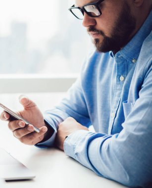 Gözlüklü yetişkin bir adamın bilgisayar masasında dizüstü bilgisayarla oturması ve gün ışığında cep telefonuyla sörf yapması.