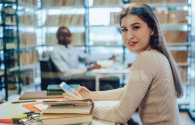 Kütüphanede oturmuş kameraya gülümserken resmi giyinmiş, akıllı telefonlara göz atan mutlu kadının yan görüntüsü.
