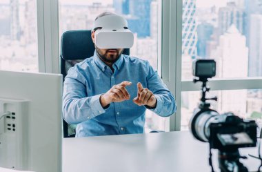 Bilgisayar masasında kameranın önünde zımbırtılarla çalışırken sanal gerçeklik hareketi yapan yetişkin bir adam.