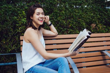 Kısa kahverengi saçlı Asyalı genç bir kadın, tebessüm ediyor ve ahşap bir bankta oturmuş, çitlere karşı kitap okuyor ve kafaya dokunurken kameraya bakıyor.