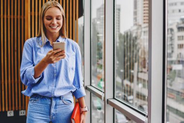 Neşeli kadın blogcu, ücretsiz 4G internetini kullanarak sosyal ağlarda modern akıllı telefon cihazında komik bildirimler okuyor. Ofis içinde cep telefonuyla sohbet eden olumlu genç kadın.